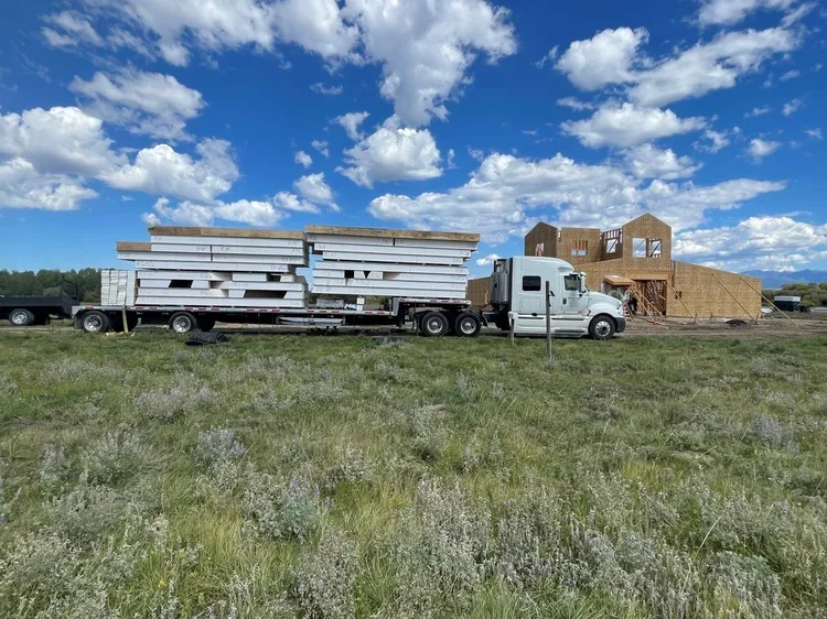 SIP wall and roof panels stacked on a flatbed truck, delivered to a home site with the shell going up in the background